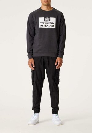 Mand iført sort sweatshirt med teksten "WEEKEND OFFENDER" og kronegrafik, sorte cargobukser og hvide sneakers, stående.