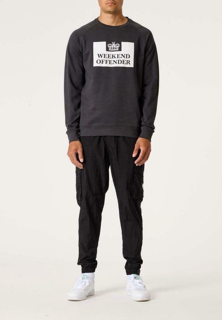Homme portant un sweat-shirt noir avec le texte "WEEKEND OFFENDER" et un graphique de couronne, pantalon cargo noir et baskets blanches, debout.