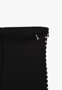 Zwarte stof met een gladde textuur, voorzien van een zijrits en witte ronde studs langs één rand. Inclusief een klein metalen accent nabij de rits.