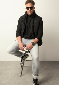 Gilet imbottito nero sopra una maglietta nera, jeans grigi chiari e sneakers nere e grigie. L'individuo indossa occhiali da sole e un orologio.