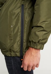 Olivengrøn puffjakke med elastiske manchetter, lynlås i siden og en lomme med trykknap, der viser en glat, skinnende stoftekstur.