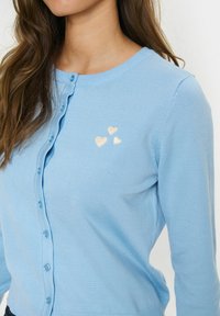 Világoskék kardigán, bordás textúrával, gombos elülső záródással, amelyen három bézs színű hímzett szív található a bal mellen.