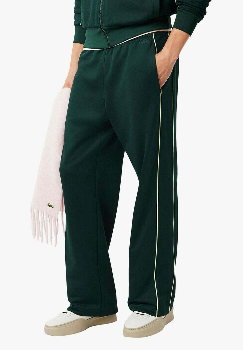 Pantaloni sportivi verdi con piping giallo a contrasto, realizzati in tessuto morbido, con una vestibilità ampia. Una sciarpa rosa chiaro è drappeggiata su un braccio.