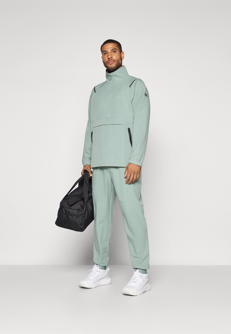 Pull-on sportif vert clair avec un col haut et des poches zippées noires, associé à un jogging assorti et des chaussures de sport blanches, tenant un sac noir.