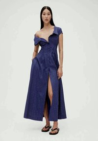 Vestido longo azul com padrão de bolinhas, design off-shoulder, fenda lateral e cintura franzida. Usado com chinelos pretos. Textura de tecido suave.