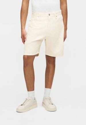 Mężczyzna w kremowych krótkich spodenkach dżinsowych, białych skarpetkach i kremowych trampkach stoi na tle jednolitego tła.