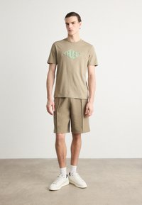 Oliven grønn bomulls t-skjorte med en lys grønn logo, kombinert med matchende shorts. Modell med hvite joggesko og sokker. Enfarget bakgrunn.