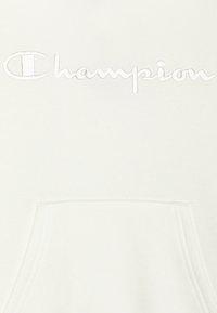 Bílá mikina s výšivkou loga "Champion" nad přední kapsou, ukazující texturu tkaniny a detaily šití.