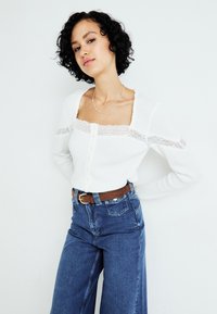 Hvit ribbestrikket cardigan med blonderkant ved halsen og pløsen, sammen med høyt liv blå denim vide bukser og et brunt belte.