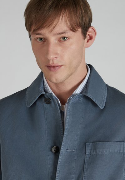 Men's blue cotton-blend jacket with a collared neckline, button closure, front pocket, and subtle texture, layered over a striped shirt.
