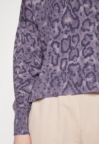 Lila Tiermuster-Pullover mit strukturiertem Strickmuster und gerippten Bündchen, kombiniert mit hellbeigen Hosen.