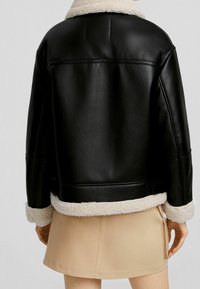 Person i svart läderjacka med vit fårskinnsfoder och beige kjol, sedd bakifrån mot en enkel bakgrund.