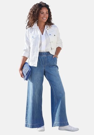 Frau trägt weiße Jacke, weißes Hemd, weit geschnittene blaue Jeans, weiße Sneaker, hält eine blaue Clutch, lächelt und steht mit einer Hand in der Tasche.