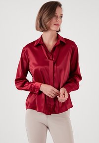 Red satin blouse with a collar and tie detail, featuring long sleeves and buttoned cuffs, displayed against a neutral background.