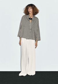 Black and white striped, oversized blazer with wide sleeves, paired with loose-fitting, white pants. Simple necklace accentuates the neckline.