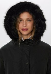 Čierna parka s kapucňou lemovanou umelou kožušinou, so zipsom a patentnými gombíkmi. Má hladkú textúru a voľný strih so bočnými vreckami.