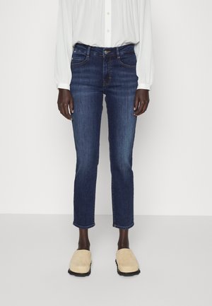 Personne portant un jean slim bleu foncé, un chemisier blanc à manches longues et des chaussures beige à enfiler, debout devant un fond blanc.