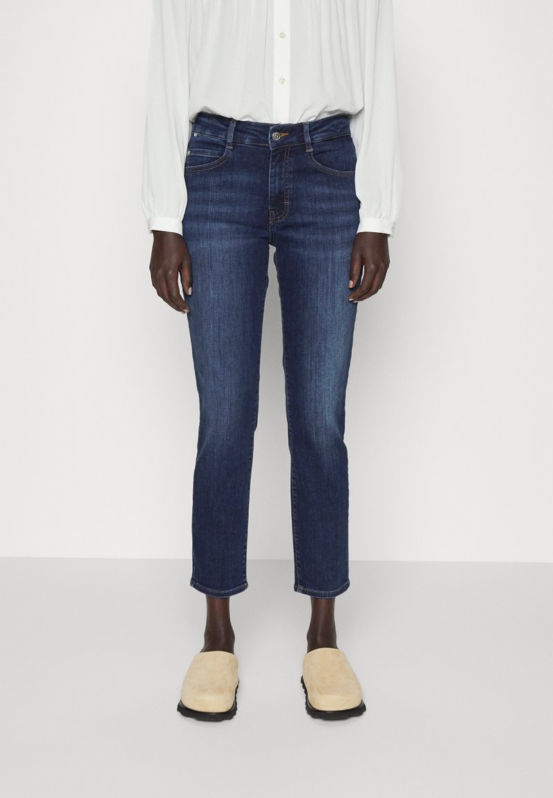 Personne portant un jean slim bleu foncé, un chemisier blanc à manches longues et des chaussures beige à enfiler, debout devant un fond blanc.