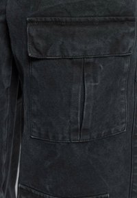 Primo piano di una tasca di una giacca di jeans nera con pattina e cuciture visibili su un tessuto testurizzato.