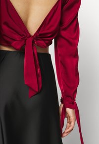 Červený saténový top s dlouhými rukávy, s hlubokým véčkovým výstřihem na zádech a uvázanými mašlemi na rukávech a na zádech, v kombinaci s černou sukní.