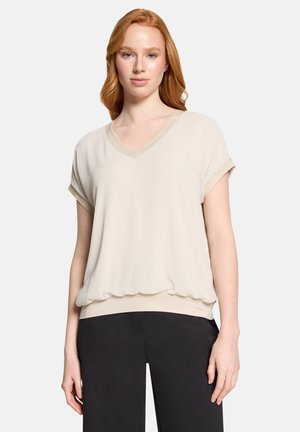 Ung kvinde med rødt hår iført en løs beige kortærmet bluse med V-hals og sorte bukser, stående mod en hvid baggrund.