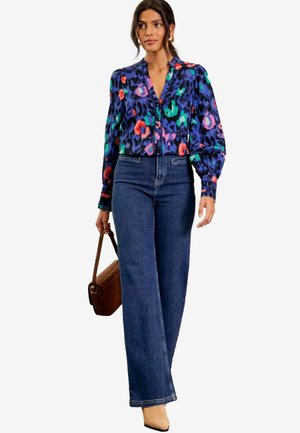 Blusa colorata a fantasia con maniche a sbuffo, abbinata a jeans a vita alta in denim blu a gamba larga. Una borsa marrone e tacchi beige completano il look.