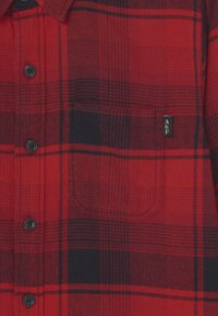 Czerwono-czarna koszula flanelowa w kratę z zapięciem na guziki, z pojedynczą kieszenią na piersi i czarnymi guzikami. Delikatna struktura widoczna.