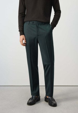 Persona que lleva pantalones oscuros a medida de color verde oscuro, mocasines negros y una camiseta de manga larga oscura, de pie con una mano en el bolsillo.
