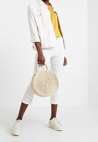 Sac rond en paille à surface texturée, porté avec un jean blanc et un haut jaune moutarde, assorti de baskets blanches.
