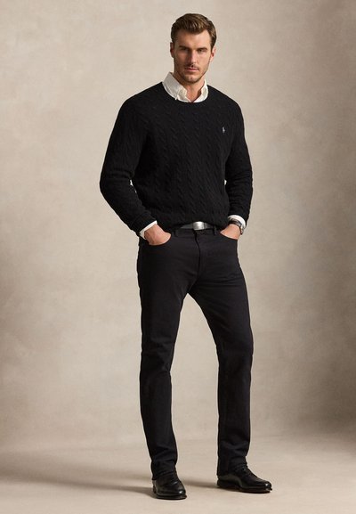 Uomo che indossa un maglione nero lavorato a trecce sopra una camicia bianca, pantaloni neri e scarpe nere, in piedi con le mani nelle tasche su sfondo uniforme.