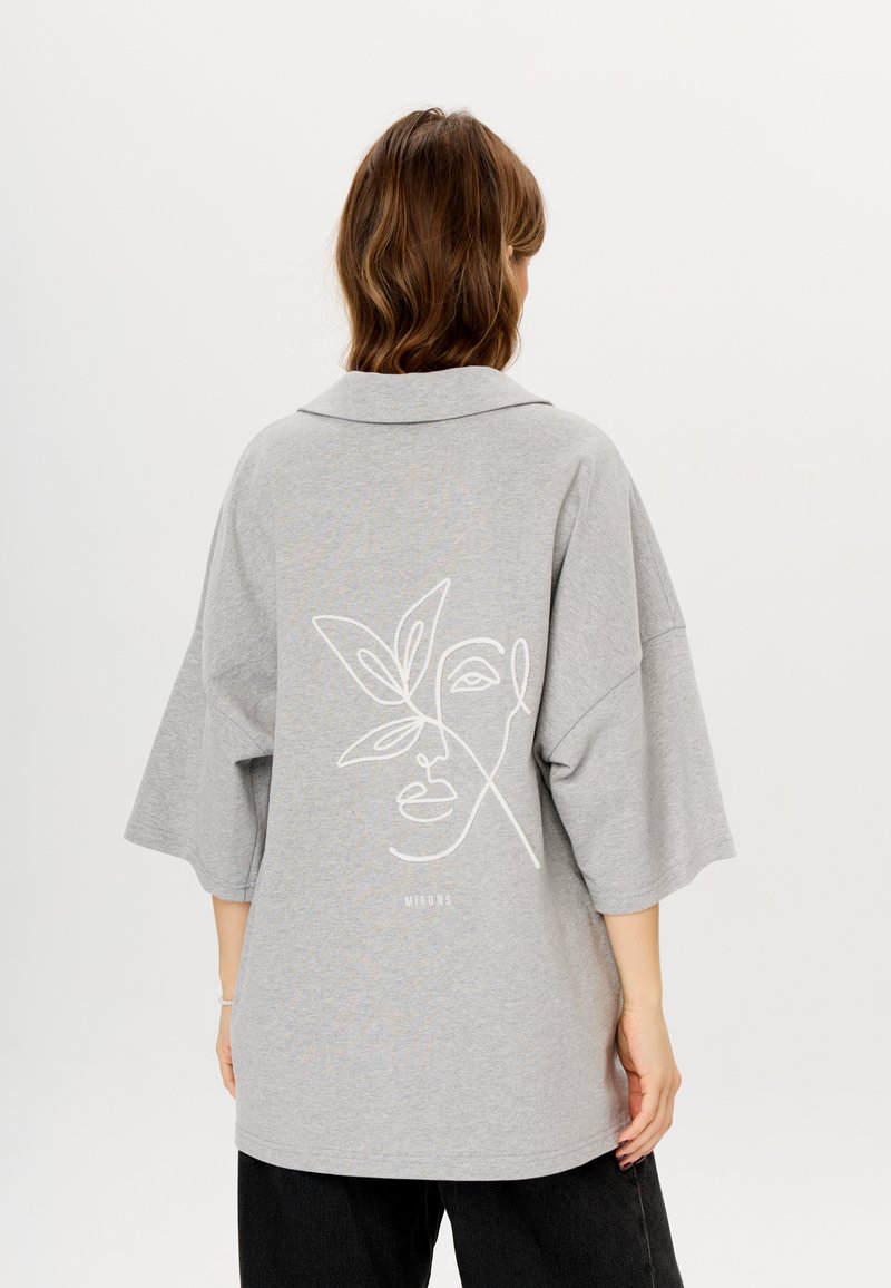 Grijze oversized shirt met korte mouwen, voorzien van een witte lijntekening van een gezicht en een bladontwerp op de achterkant. Zachte stof, ontspannen pasvorm.