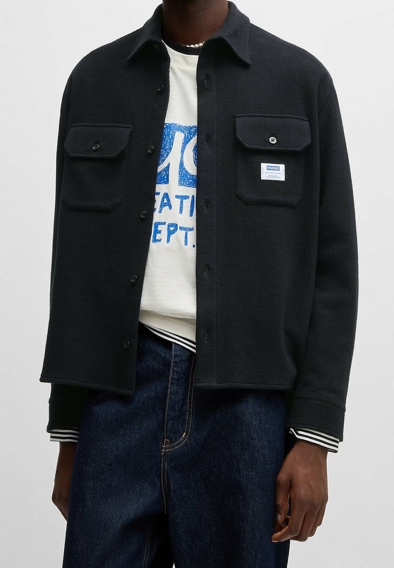 Schwarze, strukturierte Shirtjacke mit Fronttaschen, Knopfverschluss und einem Logopatch; getragen über einem weißen Grafik-T-Shirt und blauen Jeans.