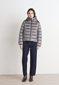 Doudoune grise avec un motif matelassé horizontal, fermeture éclair à l'avant et capuche, assortie à un jean en denim noir et des bottines brunes.