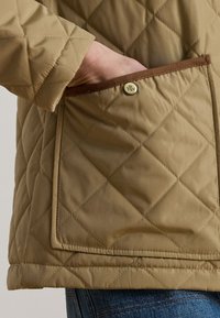 Main dans la poche d'une veste matelassée beige avec un bord marron et un bouton en métal, visible de la taille jusqu'à mi-cuisse sur un jean bleu.