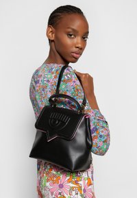 Bolso de cuero negro con forma estructurada, que presenta una solapa decorativa con un diseño de ojo en relieve y ribetes rosas. Detalles en oro.