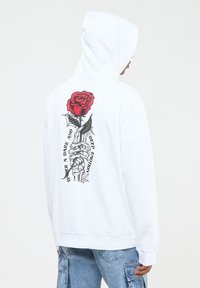 Felpa bianca con cappuccio e una grande grafica sulla schiena che presenta una rosa rossa tenuta da una mano scheletrica, circondata da testo e dettagli di ragnatela.