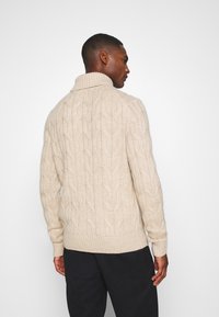 Suéter de punto beige con cuello alto, que presenta patrones texturizados y puños acanalados. Material suave y un corte relajado.
