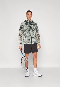 Hettejakke med glidelås i grønt og grått abstrakt mønster, brukt over svarte shorts. Holder et tennisracket og ball, med atletiske sko.