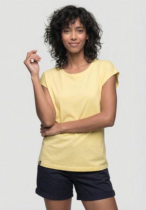 Frau mit lockigem Haar, die lächelt, trägt ein gelbes T-Shirt und dunkelblaue Shorts, posiert mit einem Arm quer über den Körper und dem anderen erhoben.