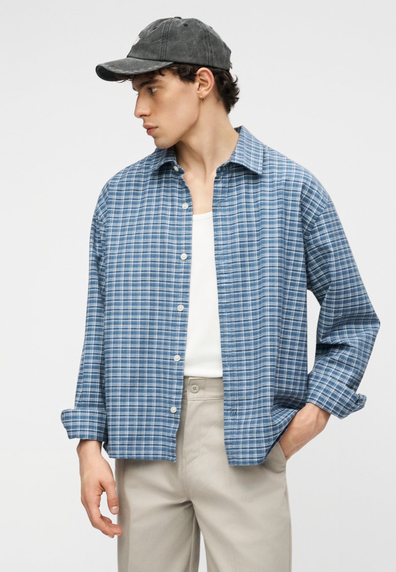 Jeune homme portant une casquette de baseball gris foncé, une chemise à carreaux bleue boutonnée sur un débardeur blanc et un pantalon beige, posant avec une main dans la poche.