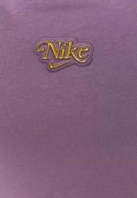 Lila Nike-plagg med ribbad textur och en guldfolierad logotyp som har varumärkesnamnet och swoosh-logotypen ovanpå.