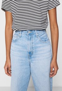 Persona vistiendo una camiseta de rayas en blanco y negro metida en unos jeans de mezclilla azul claro de cintura alta, de pie frente a un fondo liso.