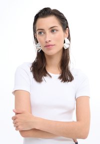 Camiseta blanca de canalé, modelo con cabello largo castaño, llevando grandes pendientes plateados reflectantes con formas geométricas.