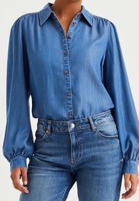 Blue denim button-up shirt with long, puffed sleeves and brown buttons, paired with high-waisted blue jeans. Smooth texture throughout.