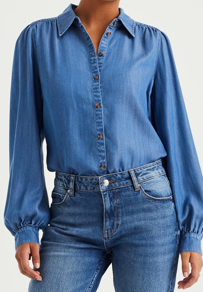 Blue denim button-up shirt with long, puffed sleeves and brown buttons, paired with high-waisted blue jeans. Smooth texture throughout.