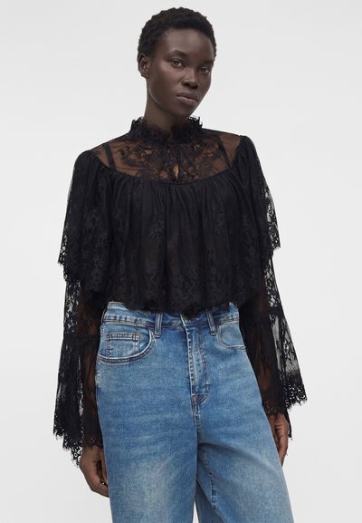 Γυναίκα με διάφανο μαύρο δαντελένιο μπλουζάκι με φαρδιά μανίκια και ψηλό λαιμό, συνδυασμένο με ψηλόμεσα μπλε τζιν.