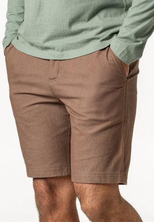Mann trägt braune knielange Shorts und ein hellgrünes Langarmshirt, die Hände in den Taschen, steht vor einem schlichten Hintergrund.