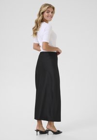 Jupe maxi en satin noir avec une texture lisse, taille haute et coupe décontractée. Associée à un haut blanc ajusté et des sandales à talons noires.