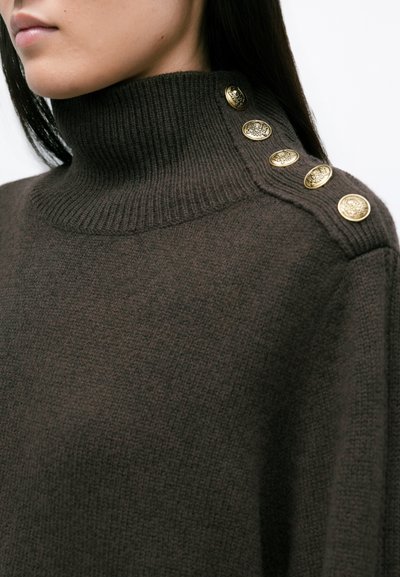 Καφέ πλεκτός σκούφος με ψηλό γιακά, διακοσμημένος με χρυσά κουμπιά κατά μήκος του ώμου, προσφέροντας απαλή υφή και χαλαρή εφαρμογή.