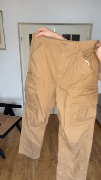 Pantalones cargo color beige sostenidos por manos, mostrando los bolsillos delanteros y el cierre de botón en una habitación con suelo de madera y muebles.